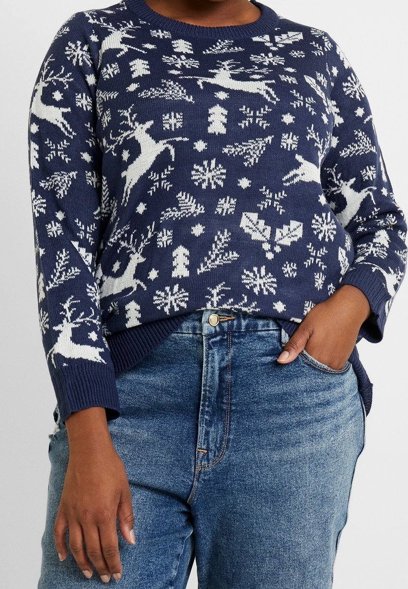 Pull bleu marine avec des motifs de rennes blancs, flocons de neige et sapins, porté avec un jean bleu, main dans la poche.