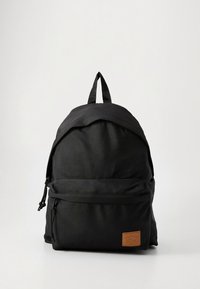Černý plátěný batoh s hladkou texturou, zakřiveným tvarem, přední kapsou na zip a malou hnědou koženou nášivkou s logem. Nastavitelné popruhy na vrchu.