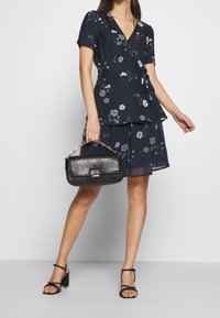 Žena v námořnických květinových šatech s kimono rukávy drží černou texturovanou kabelku, má na sobě černé sandály na blokovém podpatku a pózuje na jednobarevném šedém pozadí.