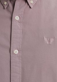 Chemise violet clair à boutons blancs avec un petit logo ailé brodé sur la poitrine près des boutons.