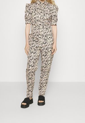 Frau in einem gemusterten, passenden Oberteil und Hosen mit Puffärmel, steht vor einem schlichten Hintergrund und trägt schwarze Plateausandalen.