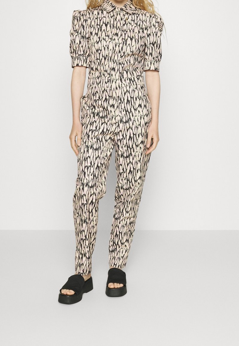 Femme portant un ensemble assorti de chemise et de pantalon à motifs avec des manches bouffantes et des sandales à plateforme noires, se tenant devant un fond uni.