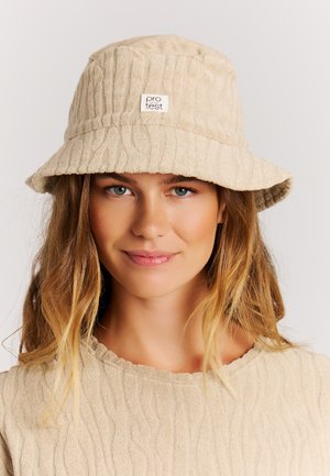 Jonge vrouw met lang blond haar, die een beige bucket hat met structuur en een bijpassende beige top met structuur draagt, zacht glimlachend.