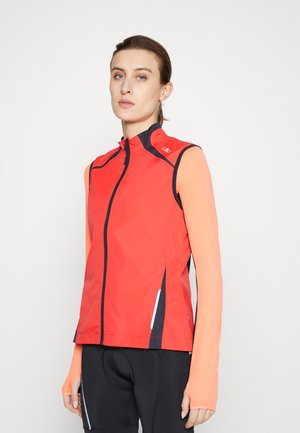 Red sleeveless jacket with a high collar, black zippers, and side accents, paired with long-sleeve orange top. Smooth, water-resistant fabric.