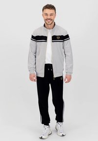 Hall hallide triibuliste ja logoga lukuga jakk, sobitatud mustade dressipükstega, millel on valged külgtriibud. Täiendatud valgete spordijalatsitega.