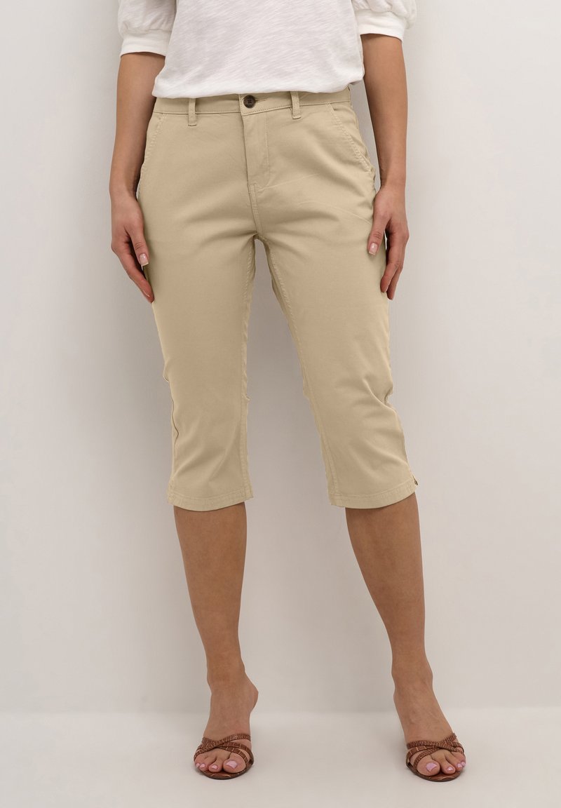 Beige Capri-Hosen aus Baumwolle mit geradem Bein, vorderen Taschen und einem Knopfverschluss. Der Saum endet direkt unter dem Knie.