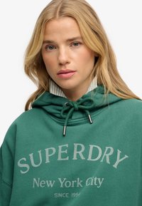 Zielona bawełniana bluza z kapturem ze sznurkiem, z paskowanym wewnętrznym kołnierzem i jasnoszarym napisem "SUPERDRY New York City". Casualowy design.