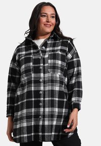 Chemise à carreaux en flanelle noire et blanche avec un col, des manches longues et une fermeture à boutons, dotée d'une poche à l'avant et d'un tissu doux et texturé.