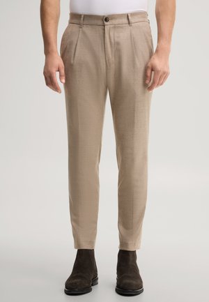 Man draagt beige op maat gemaakte broek en donkerbruine suède enkellaarsjes met een wit overhemd in de broek gestopt, staand op een effen lichte achtergrond.