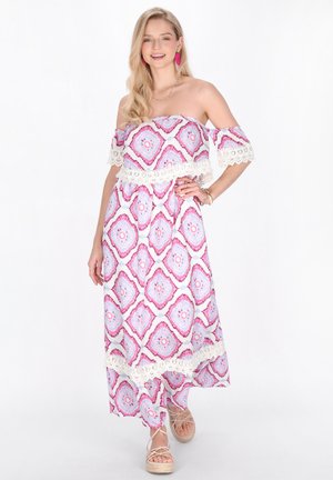 Femeie în picioare purtând o rochie maxi fără bretele, pe umeri, cu modele geometrice roz și albastre, sandale bej și cercei roz.