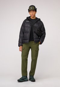 Doudoune noire, pantalon cargo vert, chemise noire, sac bandoulière noir et bottes de randonnée vertes. Matériaux texturés et design fonctionnel.