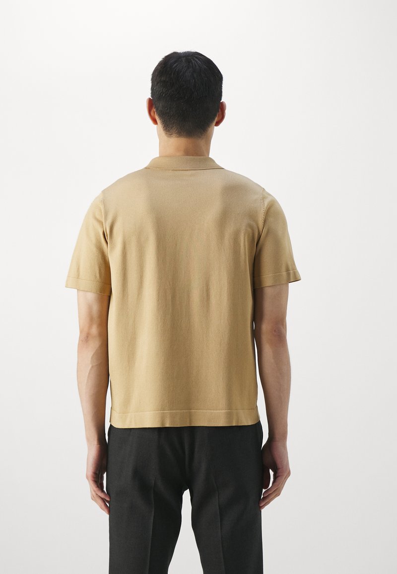 Kurzärmliges Poloshirt in Beige, aus einem glatten Stoff mit geradem Saum und klassischem Kragen. Getragen mit dunklen Hosen.