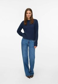 Tamno plavi pleteni džemper s dugim rukavima, uparen s svijetloplavim širokim trapericama i smeškim cipelama s potpeticama ukrašenim zakovicama.