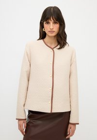 Beige quiltad jacka med ett strukturerat mönster, kontrasterande bruna detaljer och guldknappar, i kombination med en brun läderkjol.