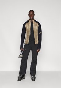 Veste beige et marine rembourrée avec fermeture éclair, couche de base noire, pantalon de ski noir et chaussures de ski ; tenant un forfait de ski dans une main.