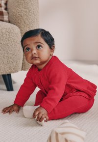 Rode gebreide trui met een kanten ontwerp, gecombineerd met bijpassende legging. De baby zit op een lichtgekleurde, gestructureerde kleed en houdt een sok vast.