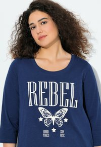 Dunkelblaues langärmliges Oberteil mit rundem Ausschnitt, das die Worte "REBEL" und eine Schmetterlingsgrafik zeigt, akzentuiert durch Sterne und Text darunter.