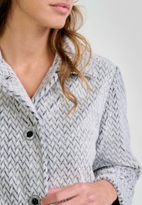 Mujer con abrigo texturizado gris claro y patrón de zigzag negro, abrochando un botón cerca del cuello, con el cabello castaño suelto.