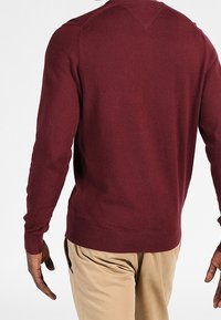 Burgunderfärgad stickad tröja med långa ärmar, ribbade mudd och nederkant, med en V-formad söm bak och slät textur. Beiga byxor syns.