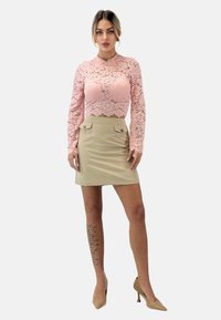 Spitzen-Crop-Top in Pastellrosa mit langen Ärmeln und hohem Kragen, kombiniert mit einem beigen Minirock mit zwei vorderen Taschen und Knöpfen.