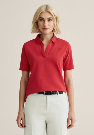 Lyshåret kvinde iført en rød kortærmet poloshirt, der er stukket ned i hvide bukser med et sort flettet bælte, står foran en ensfarvet baggrund.