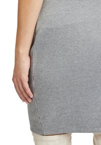 Spódnica z materiału w szaro-białą kratę houndstooth, o dopasowanym kroju. Widoczny szew boczny i gładka faktura. Ręka oparta na boku spódnicy.