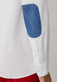 Chemise blanche avec des coudières en denim et des coutures contrastées. La chemise présente des poignets à boutons et un ourlet arrondi.