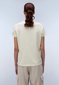 Maglietta leggera beige con maniche corte e collo rotondo. Il tessuto sembra morbido con una texture liscia. Presenta un piccolo logo sulla manica.