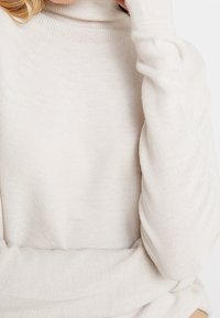 Pull à col roulé beige clair en mailles texturées. Il présente des manches longues et une coupe décontractée. Design minimaliste sans embellissements.