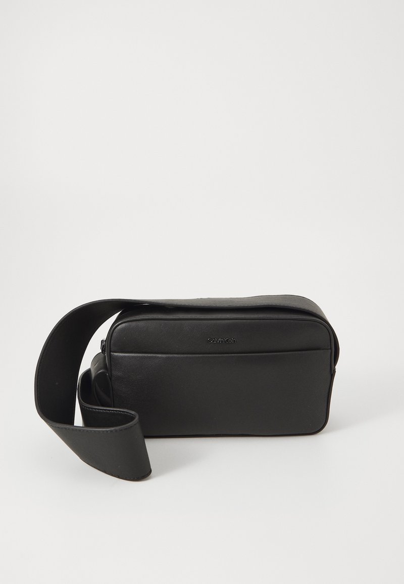 Bolso cruzado de cuero negro con forma rectangular, cierre de cremallera y correa ajustable. Presenta un sutil logo de la marca en la parte frontal.