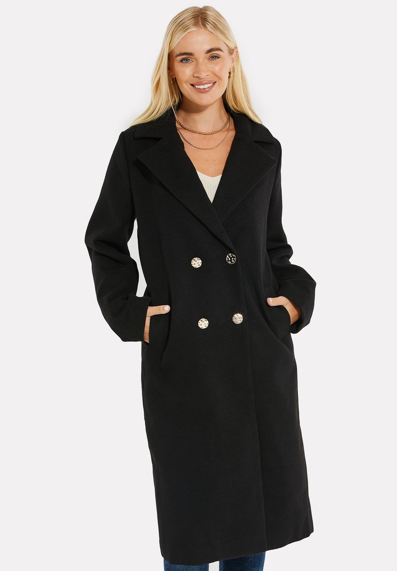 Cappotto nero doppiopetto con grandi bottoni metallici, grandi revers e tasche laterali. Realizzato in un tessuto morbido con una consistenza liscia.