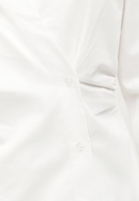 Close-up van gerimpelde witte blousestof met twee doorzichtige knopen aan de voorkant en lichte plooien aan de zijkant.