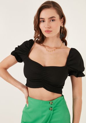 Black cropped top with puffed sleeves and ruched detailing; paired with a green skirt featuring button accents. Gold necklace completes the look.