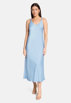 Frau trägt ein ärmelloses, hellblaues Midikleid mit dünnen Trägern und nudefarbene offene Zehentheels, steht vor einem weißen Hintergrund.