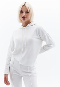 Sudadera blanca de punto con un bolsillo delantero, cordón ajustable y corte relajado, combinada con pantalones blancos a juego con costuras en los laterales. Textura suave.