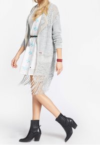 Hellgraue gehäkelte Strickjacke mit Fransen am Saum, getragen über einem weißen Blumenkleid, kombiniert mit schwarzen Stiefeletten mit Blockabsatz.