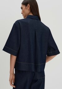 Navy denim short-sleeve shirt with a collared neck, visible seams in contrasting yellow thread, and a relaxed fit with a back pleat.