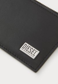 Čierna kožená peňaženka s textúrovaným povrchom, prešívanými okrajmi a striebornou kovovou logou, ktorá hovorí "DIESEL PRE ÚSPEŠNÝ ŽIVOT."