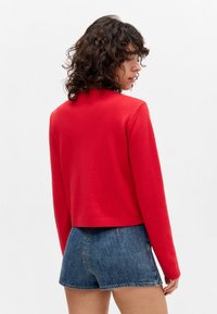 Red knitted sweater with a high neckline and ribbed hem, paired with denim shorts. Texture appears soft and smooth.