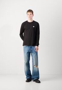 Zwarte lange mouwen shirt met een klein logo, gecombineerd met lichtblauwe distressed jeans met scheuren. Model staat voor een egale witte achtergrond.