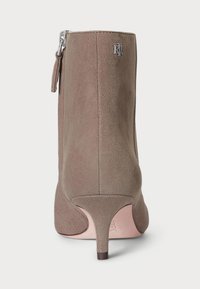 Taupe velúr bokacsizma elegáns dizájnnal, ezüst logóval, oldalsó cipzárral és keskeny sarkú kivitelezéssel. Sima textúra, minimális részletezés.