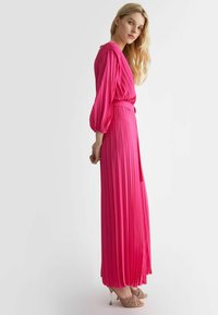 Abito rosa plissettato con maniche lunghe e puffate e vita aderente. Lunghezza fino al pavimento, con una texture liscia e una finitura lucida, abbinato a tacchi leggeri.