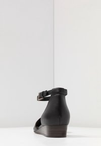 Chaussure compensée en cuir noir avec un dos fermé, une lanière de cheville et un talon en bois. Design élégant et texture lisse.