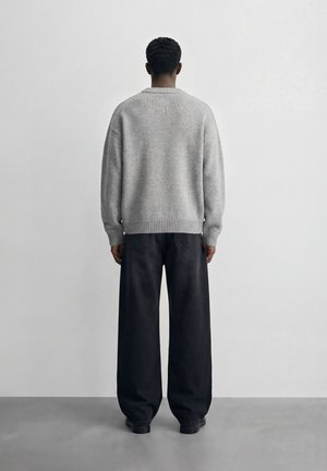 Osoba stojąca twarzą do ściany, ubrana w jasnoszary sweter i szerokie czarne spodnie na szarej podłodze i białym tle.