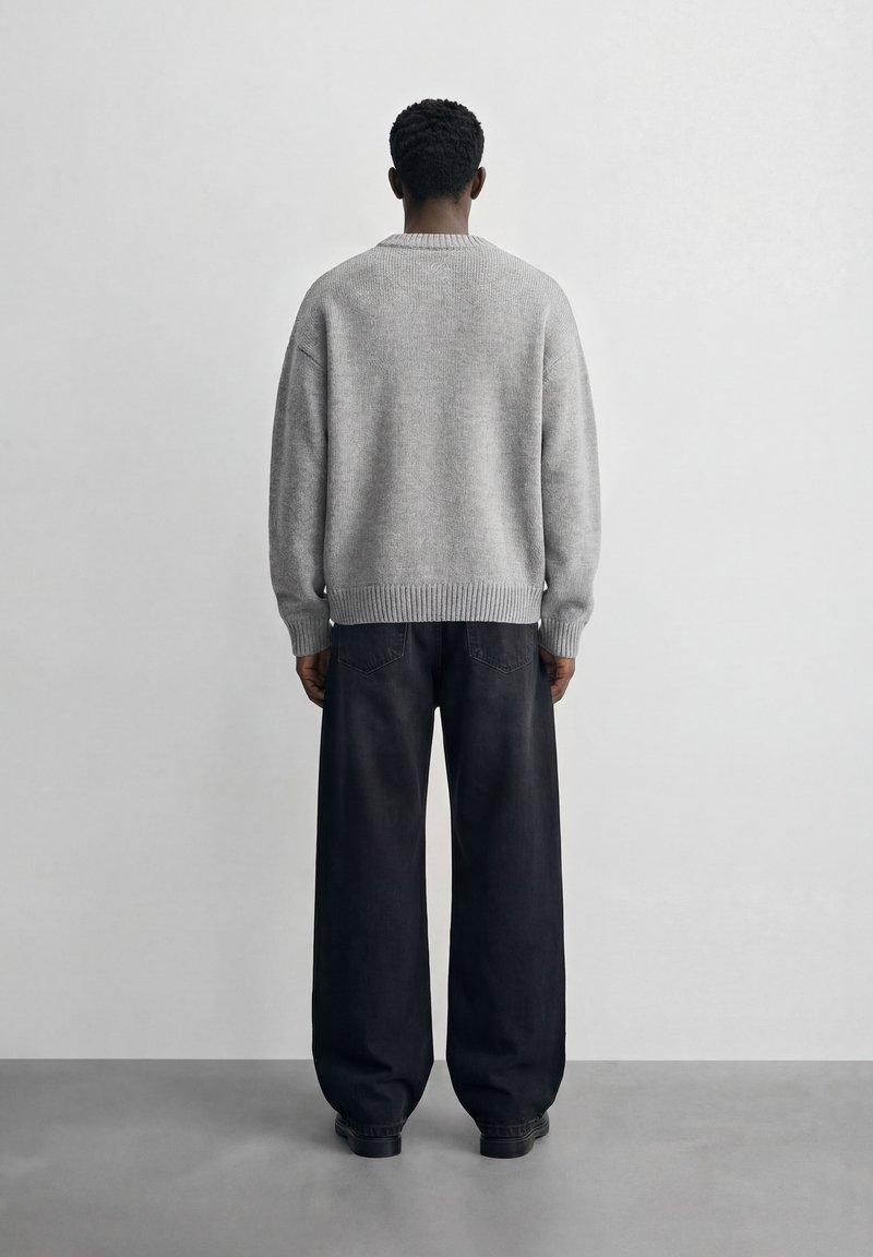 Άτομο που στέκεται στραμμένο προς τον τοίχο, φοράει ανοιχτό γκρι πουλόβερ και μαύρο παντελόνι με φαρδιά πόδια, σε γκρι πάτωμα και λευκό φόντο.