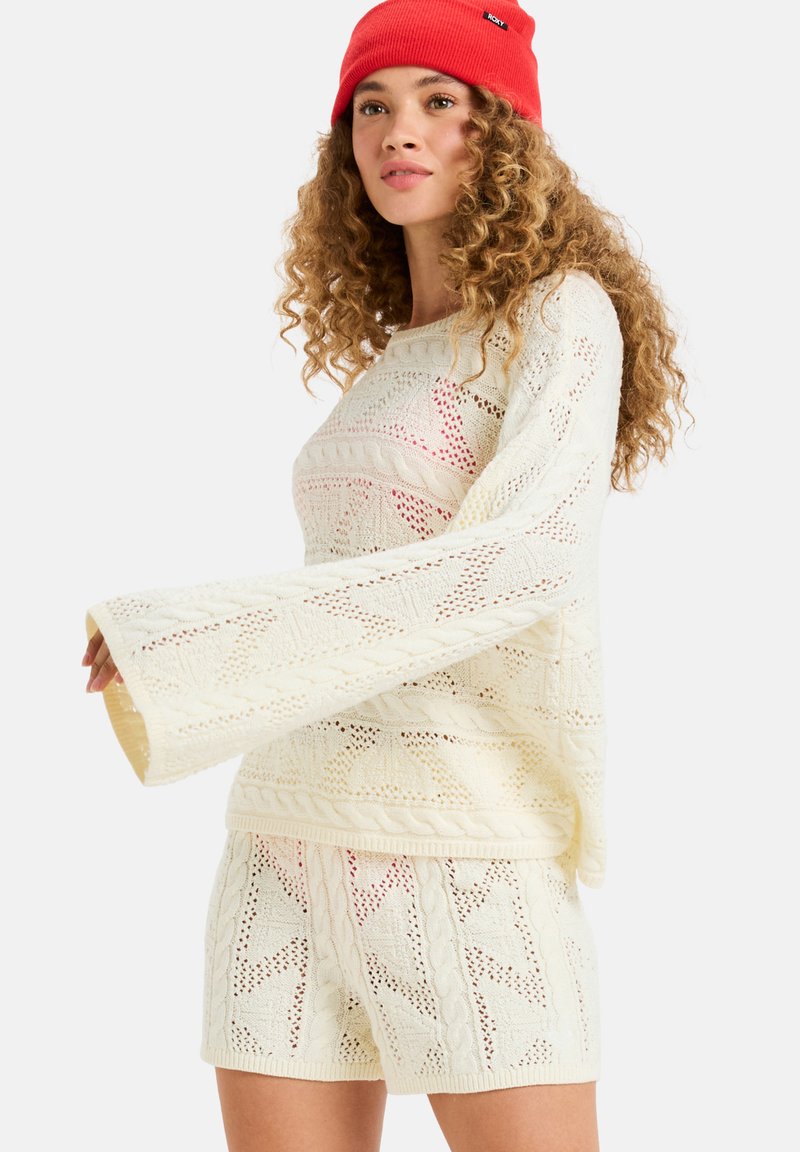 Sweter i szorty w kolorze kremowym, z otwartymi wzorami i dzwonowymi rękawami. Zestaw zawiera czerwoną czapkę jako akcent.