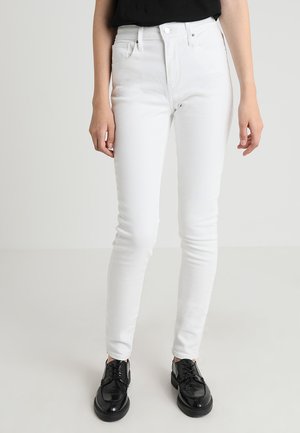 Jeans skinny de denim blanco con cintura alta, que cuentan con cinco bolsillos, un cierre de botón y una textura suave. Combinados con zapatos negros con cordones.