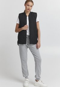 Schwarze gesteppte ärmellose Weste mit hohem Kragen, getragen über einem weißen T-Shirt und grauen Sweatpants, vor einem neutralen Hintergrund.