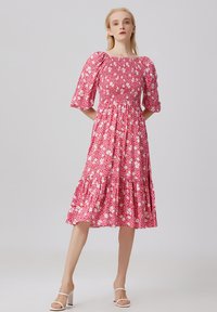 Robe florale avec un col carré, des manches bouffantes et un corsage smocké. Fond rouge avec des fleurs blanches et roses. Longueur genoux avec des volants.