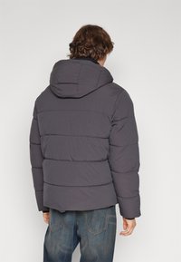 Grå puffsjakke med hætte, der har et quiltet design, elastiske manchetter og en afslappet pasform. Materialet ser glat og isoleret ud.